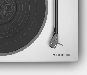 Detail shot of the lower right corner of a turntable, showing the black tonearm, stylus, and a portion of the circular platter, with the Cambridge logo displayed on the minimalist, light-colored plinth.