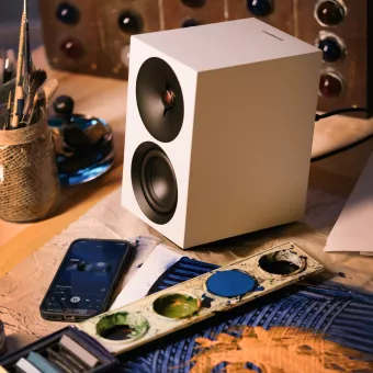 White speaker on a cluttered artist's table with paints, brushes, and a smartphone.