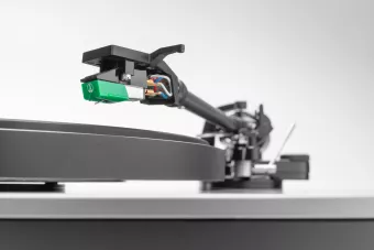 Macro side view of a turntable’s black tonearm and green cartridge, highlighting the exposed colored wiring and fine stylus positioned above a matte black platter and silver plinth.