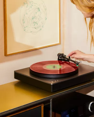 Wide shot of a modern living space with a person placing the tonearm onto a red vinyl record on a sleek black turntable, surrounded by framed artwork and hi-fi equipment.